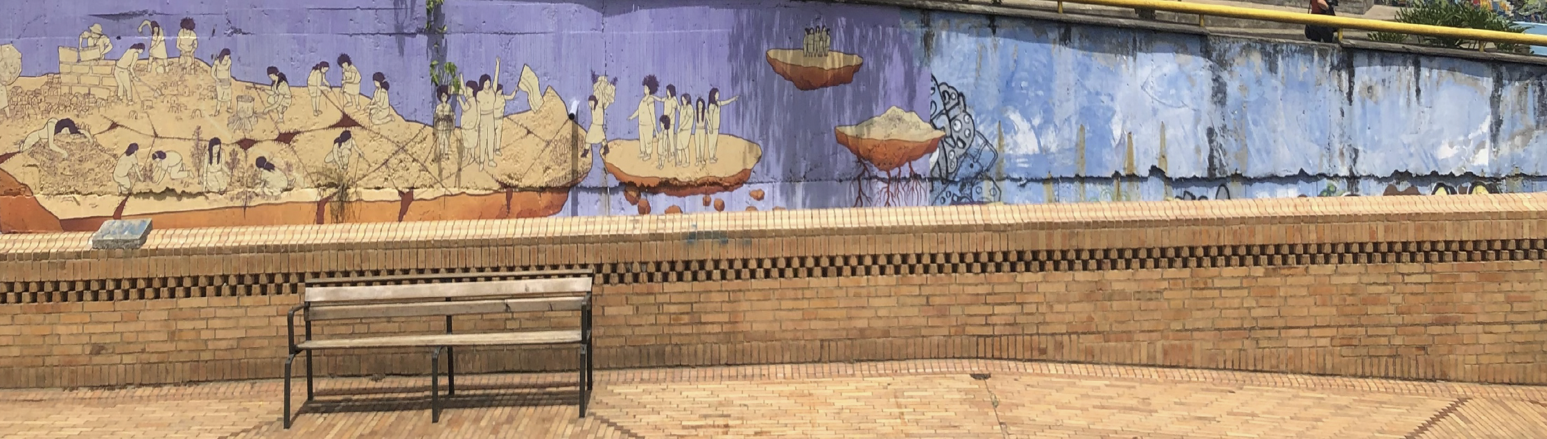 Panoramafoto einer bemalten Mauer über einer Backsteinbank: Das Wandgemälde zeigt mehrere schwebende Inseln vor violett-blauem Hintergrund, auf denen stilisierte Menschen gemeinsam arbeiten, graben und diskutieren. Einige Inseln haben sichtbare Wurzeln, im Vordergrund stehen eine Holzbank mit Metallgestell und ein gepflasterter Platz aus Backsteinen.