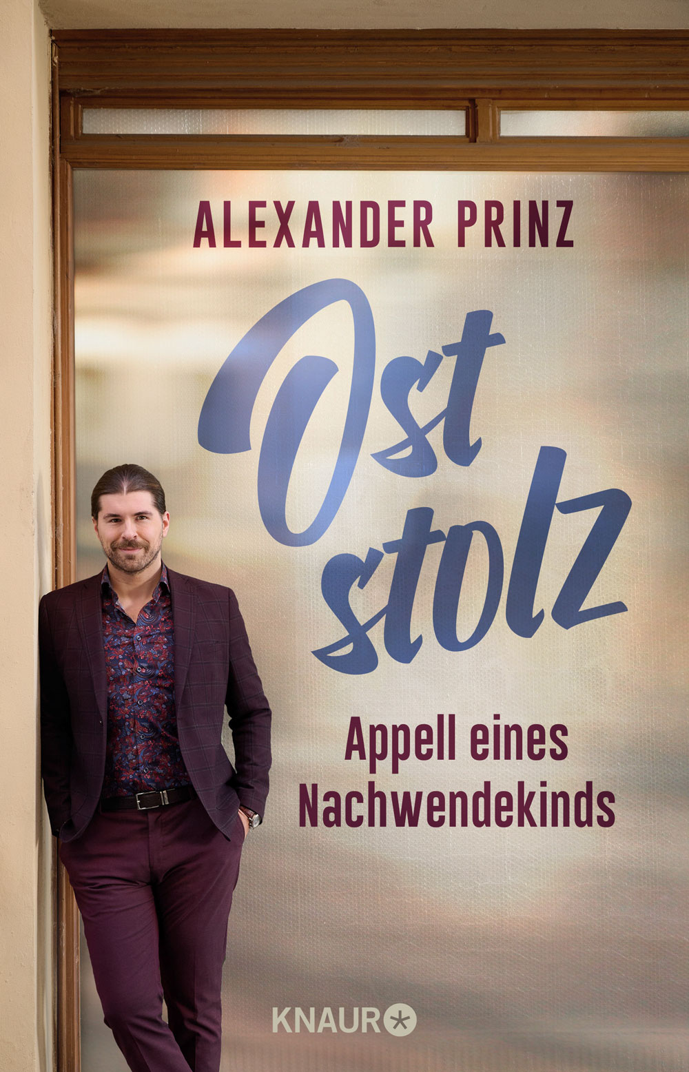 Buchcover „Oststolz – Appell eines Nachwende­kinds“ von Alexander Prinz: Eine Person in bordeauxfarbener Anzughose und Sakko lehnt lässig an einer Hauswand vor der groß gesetzten Titeltypografie, unten das Verlagslogo Knaur.