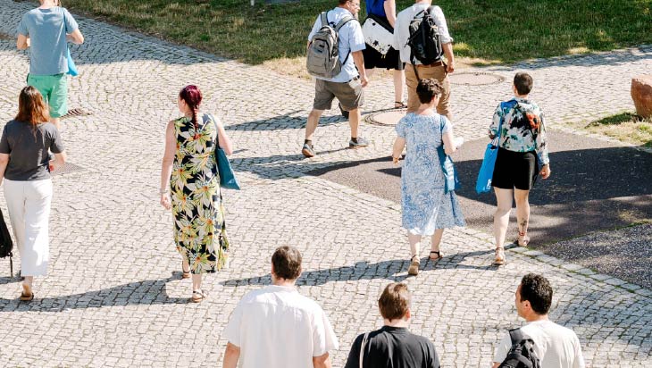 Mehrere Personen gehen über einen hell gepflasterten Weg auf einem Campusgelände. Einige tragen Rucksäcke oder blaue Stofftaschen, im Hintergrund sind Rasenflächen und weitere Wege zu sehen. Die Aufnahme zeigt die Personen überwiegend von hinten oder der Seite.