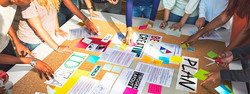 Mehrere Personen stehen um einen Tisch und arbeiten gemeinsam an einem kreativen Projekt. Auf der Tischfläche liegen zahlreiche bunte Haftnotizen, Ausdrucke und Plakate mit Worten wie „IDEA“, „PLAN“ und „BE CREATIVE“. Die Hände der Beteiligten zeigen auf verschiedene Materialien oder beschriften Zettel, was eine kollaborative Brainstorming- oder Planungsphase in einem Team widerspiegelt.