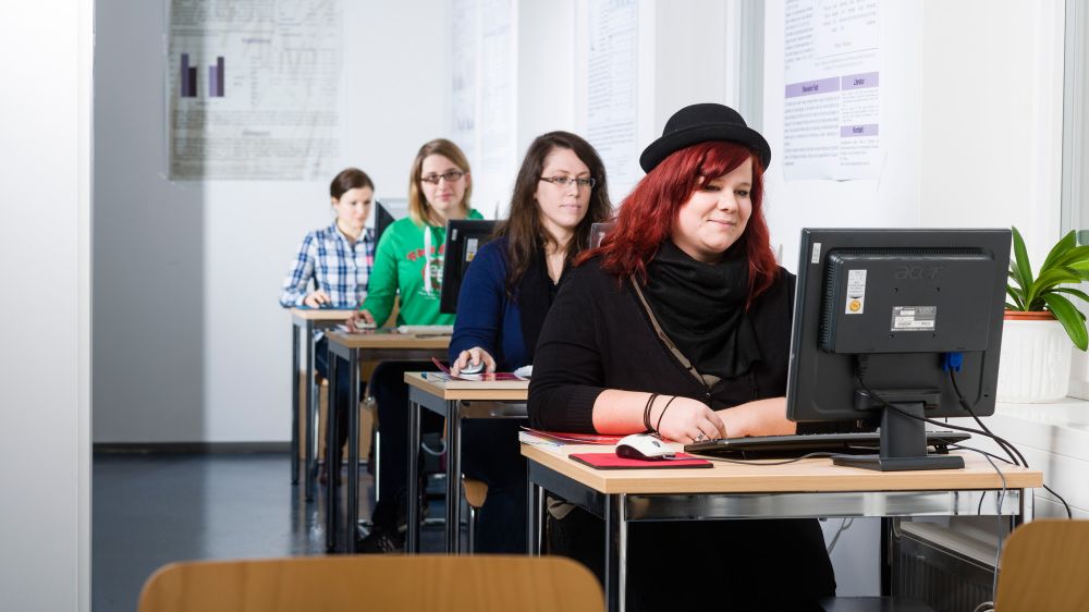 vier Studierende sitzen hintereinander an kleinen Tischen mit PC und Monitor